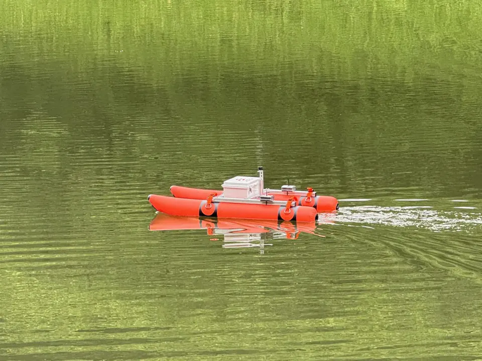 緑色の水面に浮かぶ、オレンジ色の浮き袋を2本並べた小型の無人ボート。中央に機材の箱とセンサーが載っている