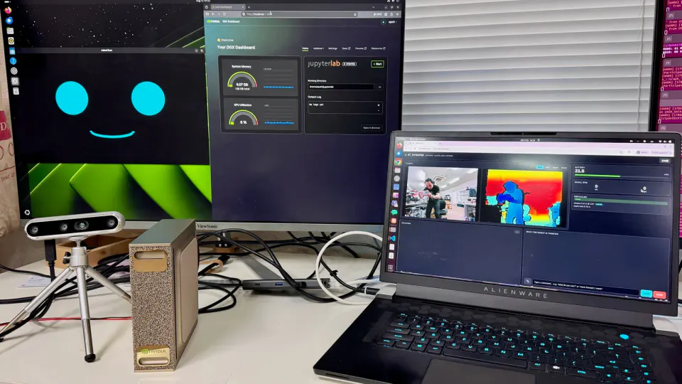 机の上に小型のAIコンピューターとカメラ、ノートパソコンが並ぶ。奥のモニターには、青い目のような顔の表示と、AIの処理画面が映っている