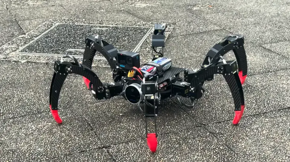 屋外のタイルの上に立つ、黒い6本脚のロボット。脚の先は赤く、胴体の中央にカメラが付いている