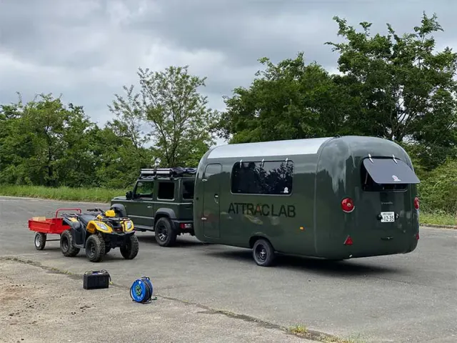 舗装された広場に止まる、ATTRAC LABと書かれた深緑色のトレーラーと四輪駆動車。手前に小型の四輪バギーと赤い荷車が並び、奥に木々が茂っている